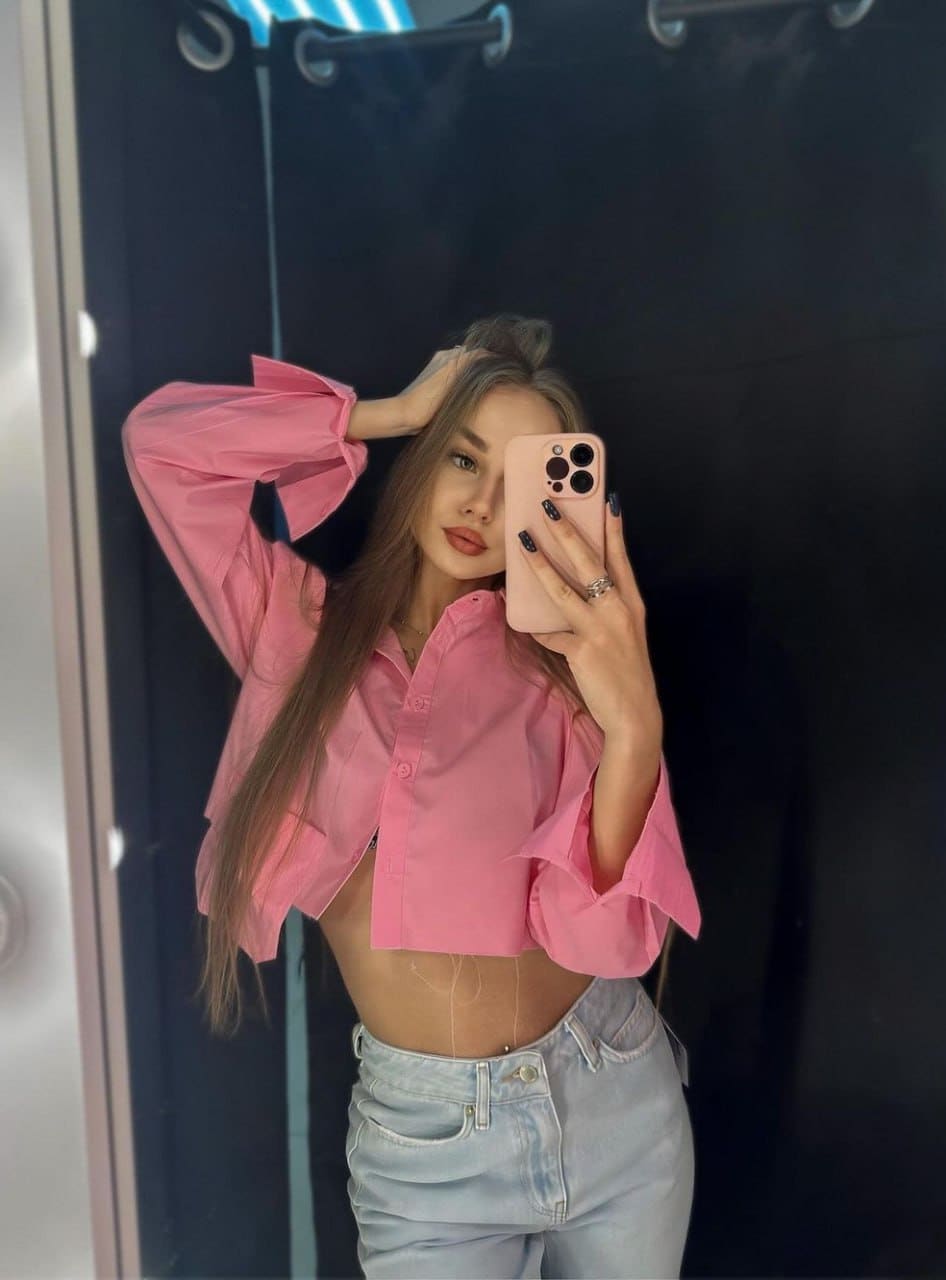 A person with long hair takes a mirror selfie in a pink cropped button-down shirt and light blue jeans.