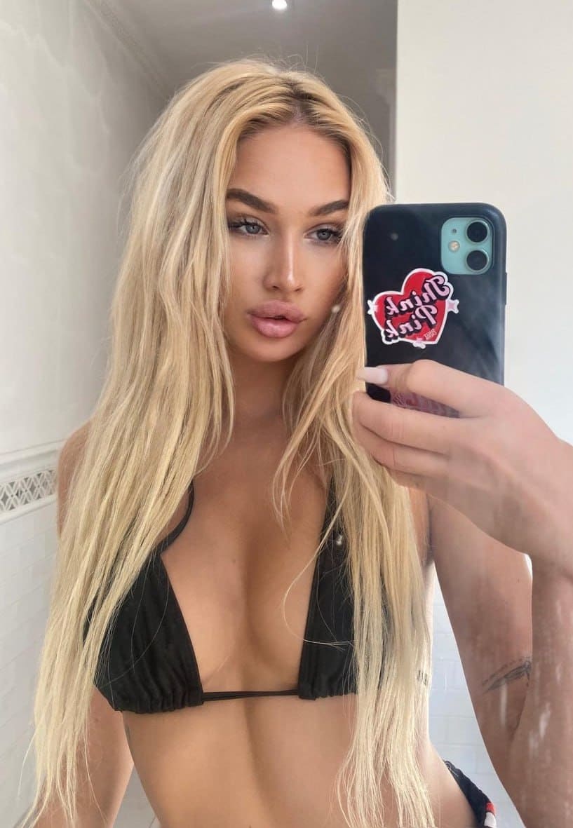 Mirror selfie showing a person with long blonde hair in a black bikini top, holding a phone with a heart-shaped sticker.