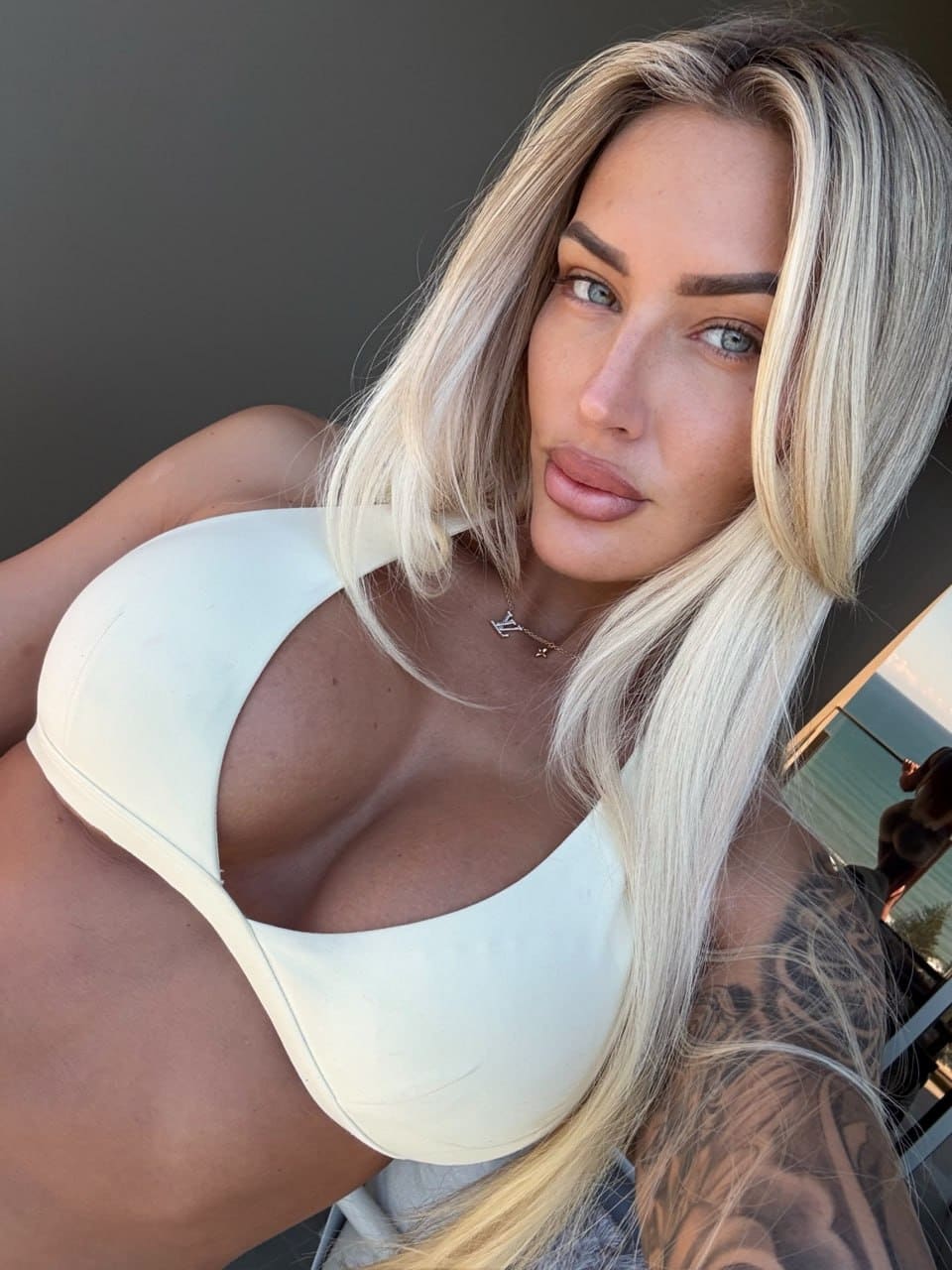 Anika with long blonde hair and a tattooed arm poses in a white one-piece swimsuit with a deep neckline against a neutral background.