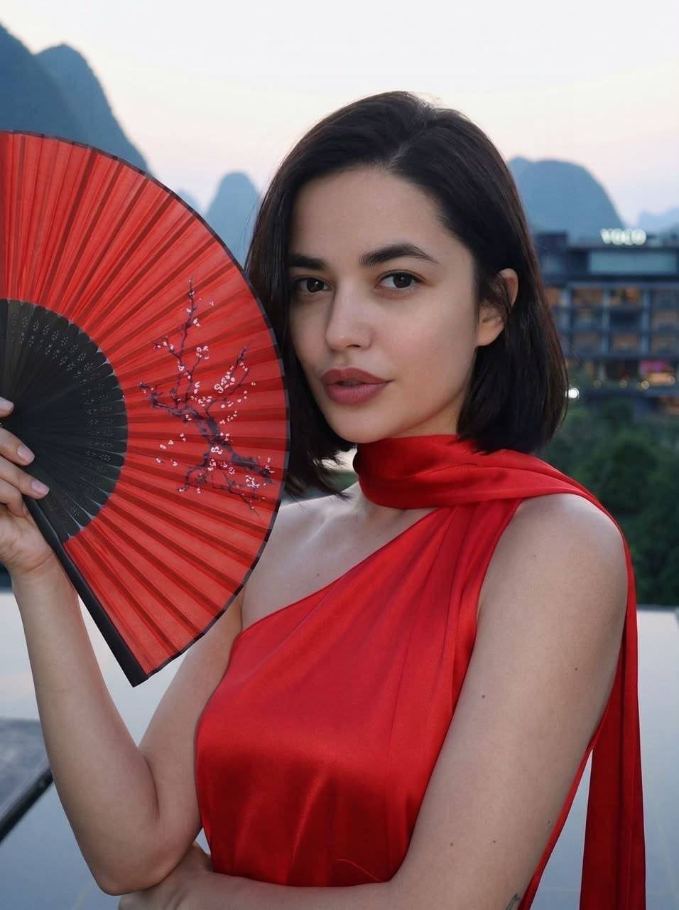 Lilian stands outdoors in a red one-shoulder dress holding a fan, with mountains and a building in the background.