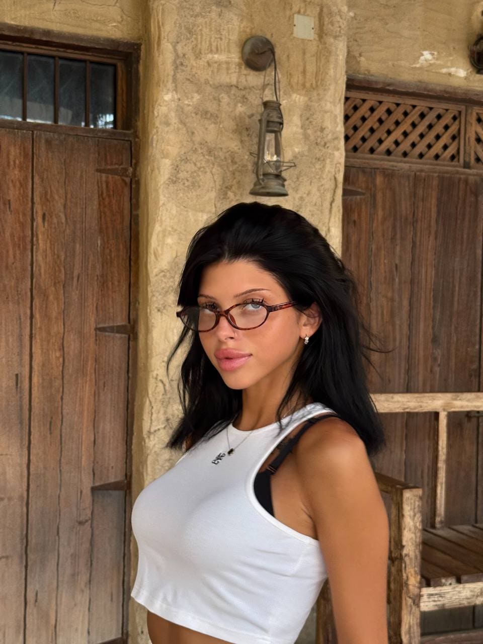 Lika stands in a white tank top exposing her midriff before a rustic stone wall and wooden door.