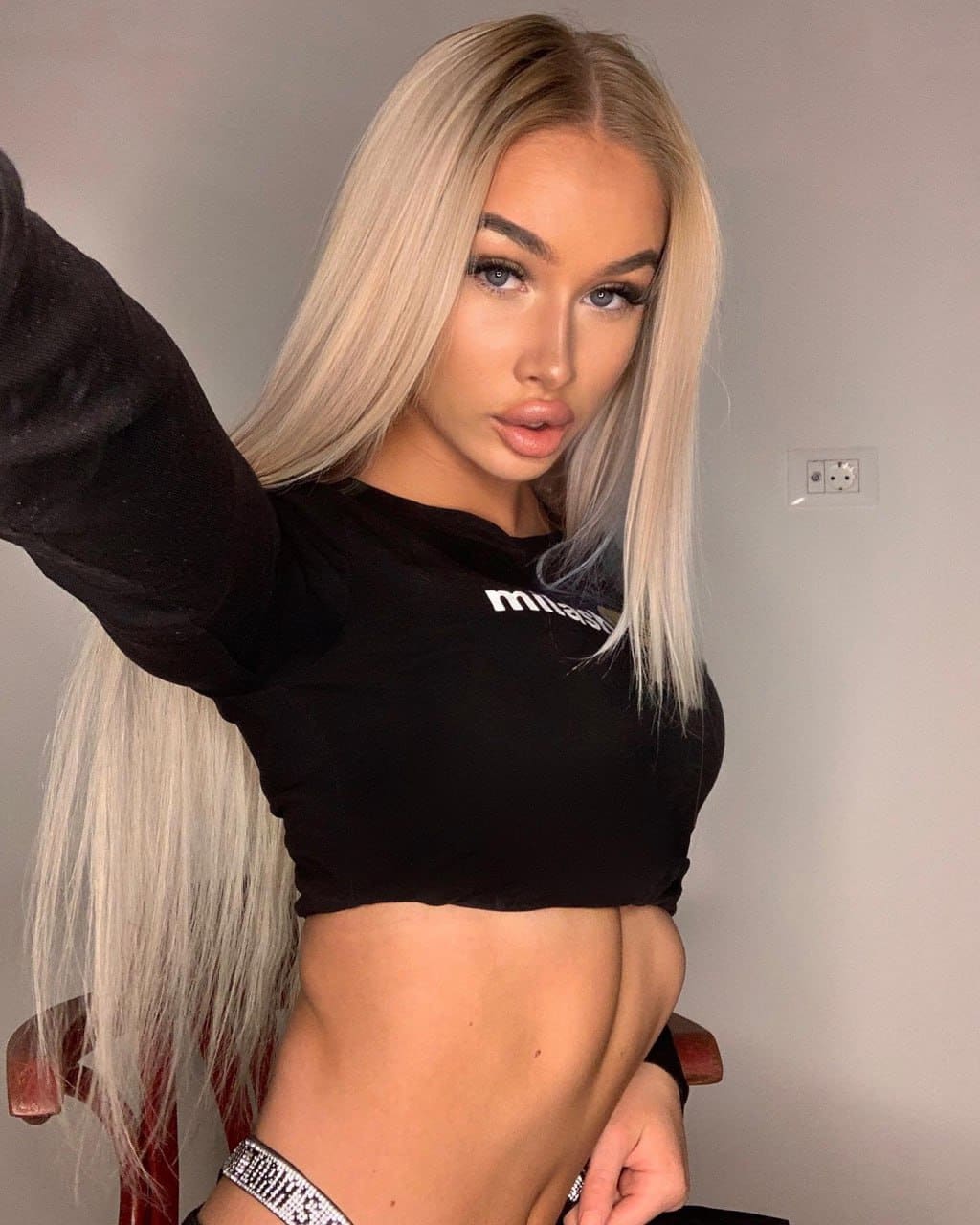 Polina stands against a plain wall with one arm raised, wearing a black long-sleeved crop top and a sparkly waist belt.