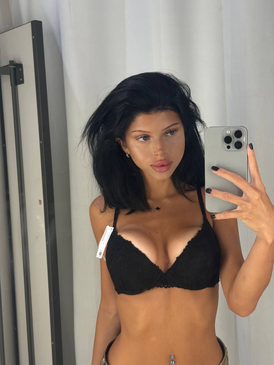 A person takes a mirror selfie in a black lace bra and low-rise bottoms against white curtains.