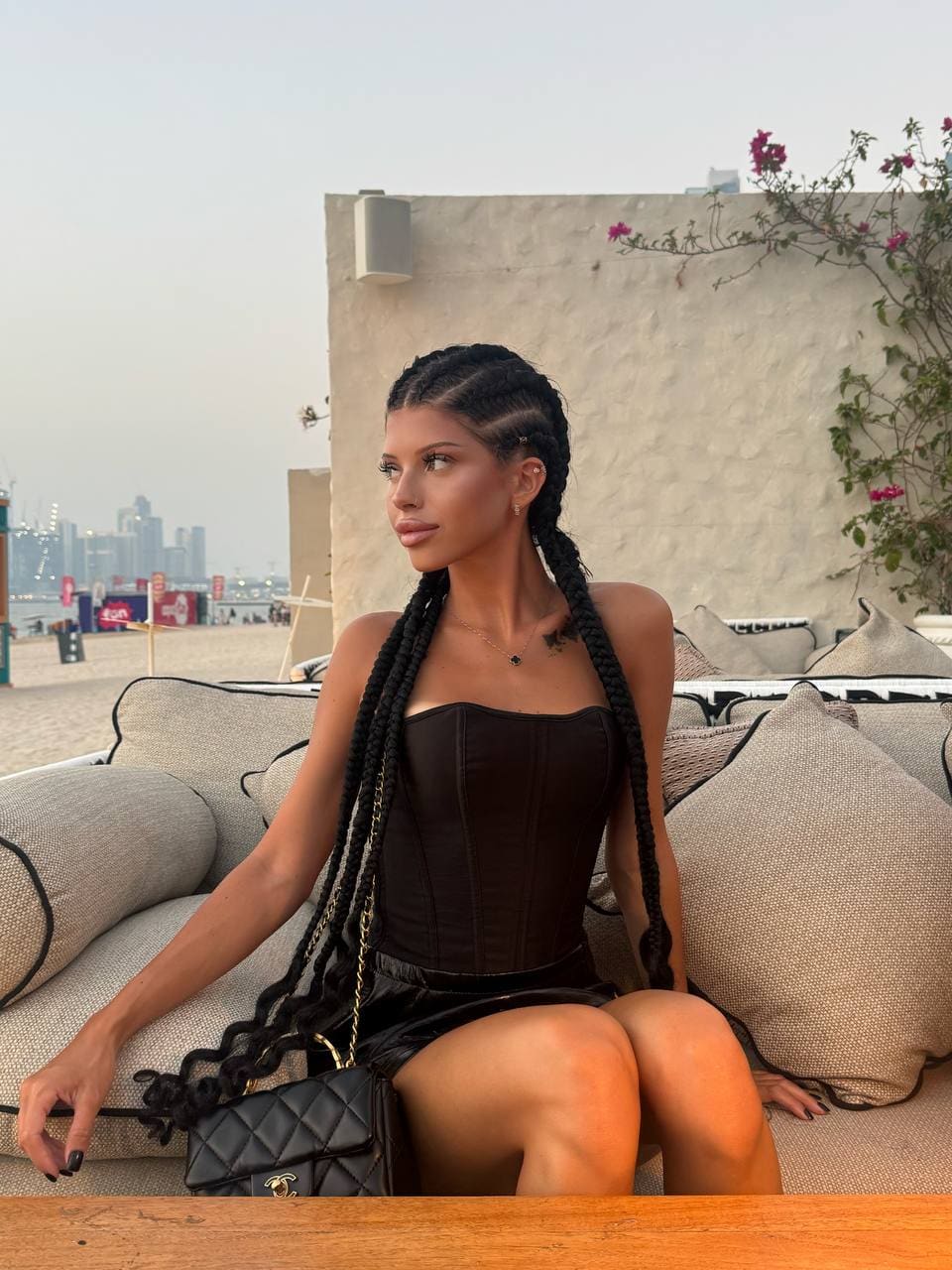 Seated on a beige sofa, a woman in a black corset top and skirt holds a quilted bag against a city backdrop.