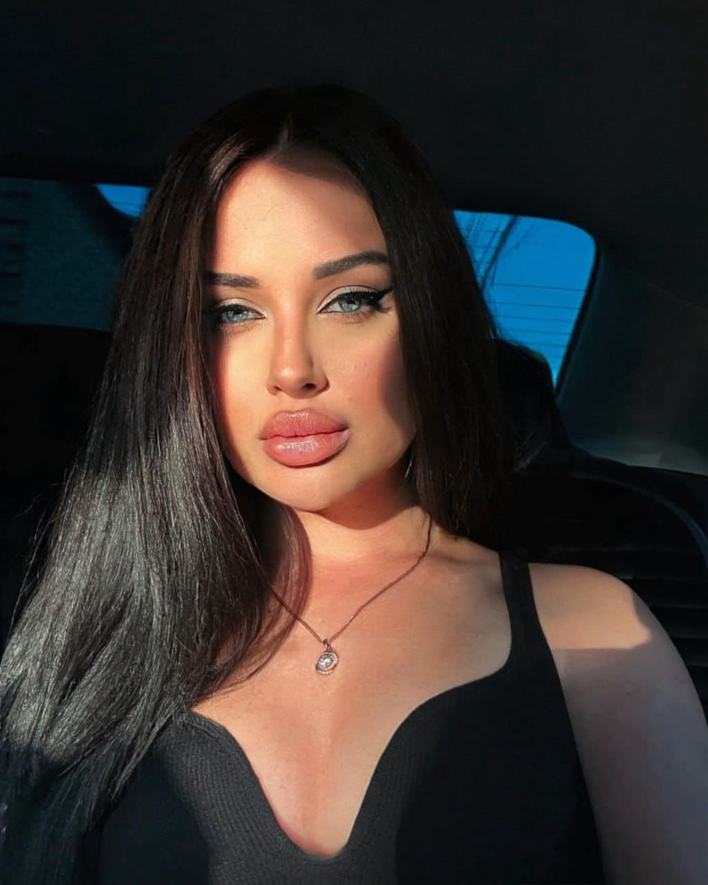 A woman with winged eyeliner sits inside a vehicle wearing a black tank top and necklace.