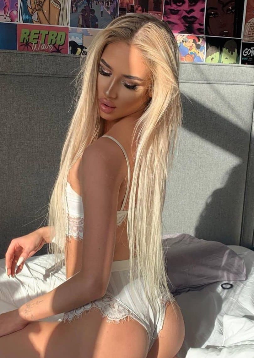 A person with long blonde hair sits on a bed in white lace lingerie, posing with their back partially turned.