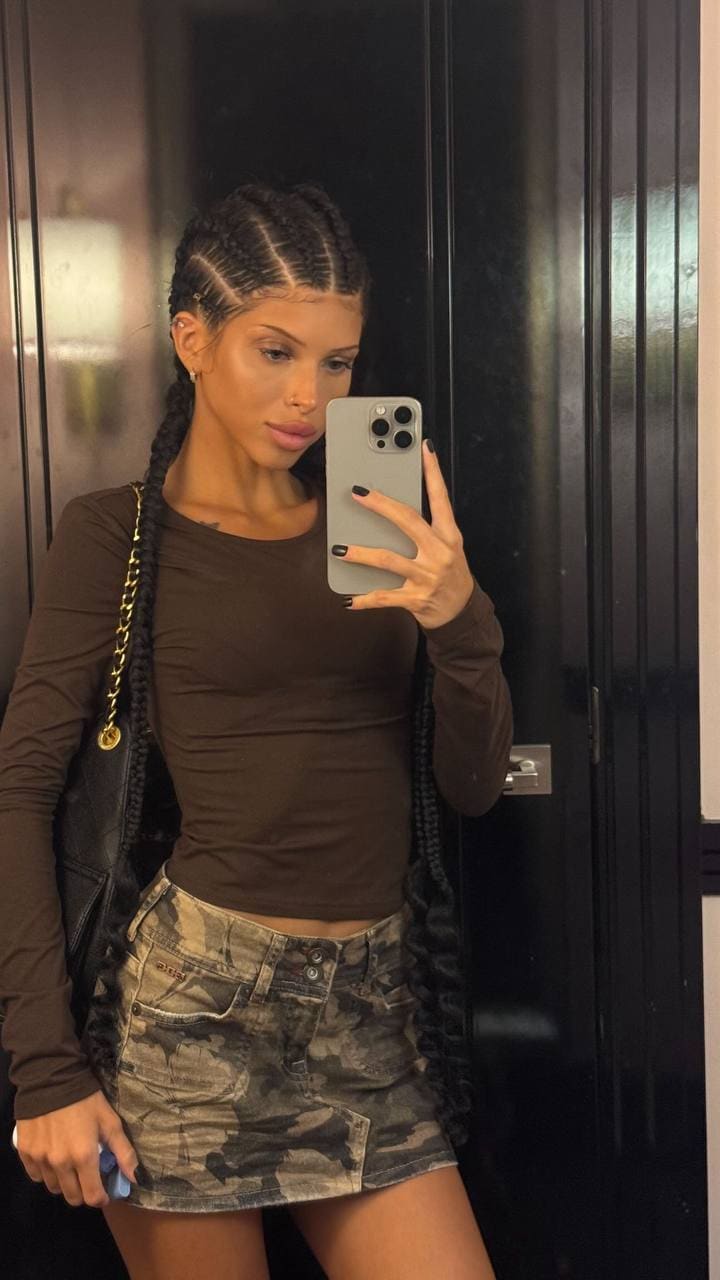 A person with braided hair takes a mirror selfie wearing a brown long-sleeved top and camouflage mini skirt.