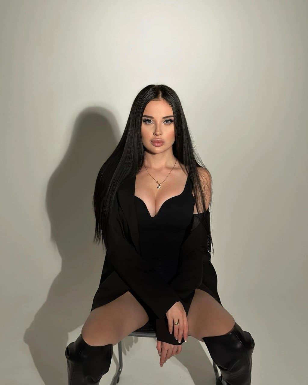 A woman sits on a chair in a black dress with bare shoulders and thigh-high black boots.