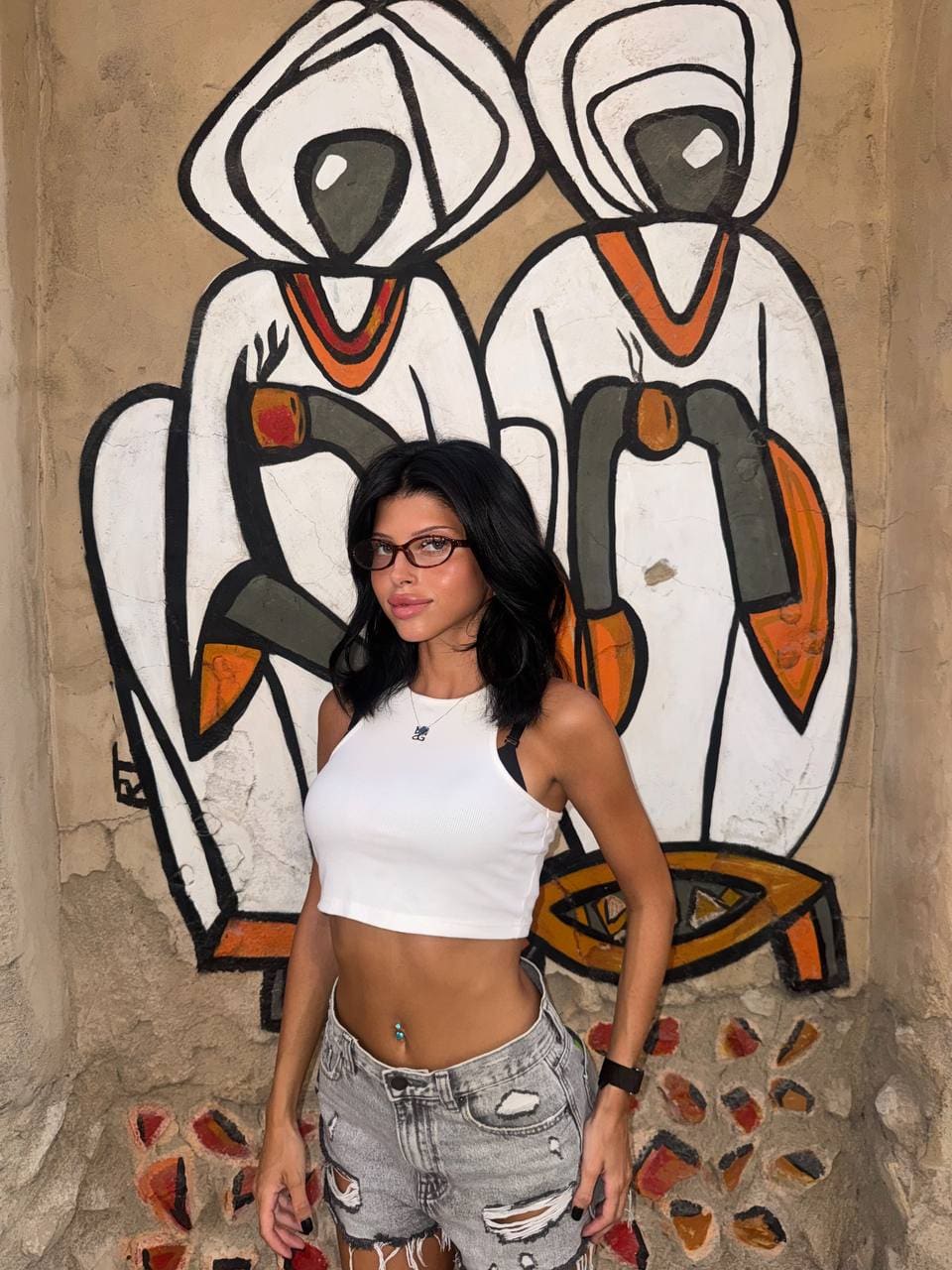 A woman in a white ribbed crop top and distressed denim shorts stands before a wall mural.
