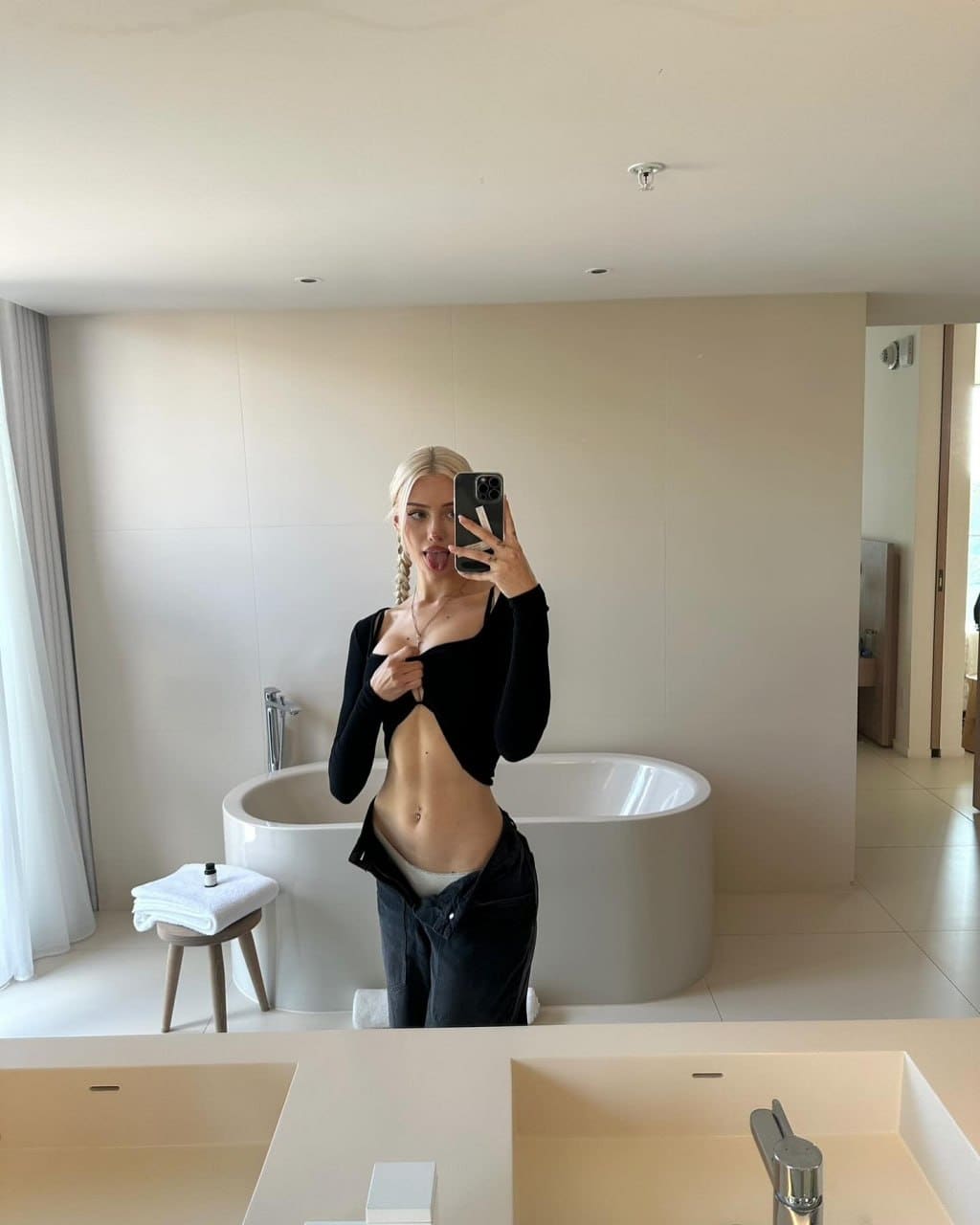 A person with blonde braids takes a mirror selfie in a bathroom, wearing a black top with a midriff cutout and dark pants pulled down.