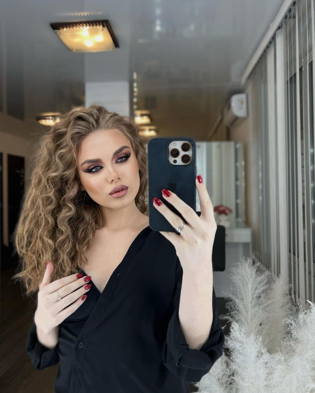 Mirror selfie of a woman in a black V-neck shirt, standing near vertical blinds and white plants.