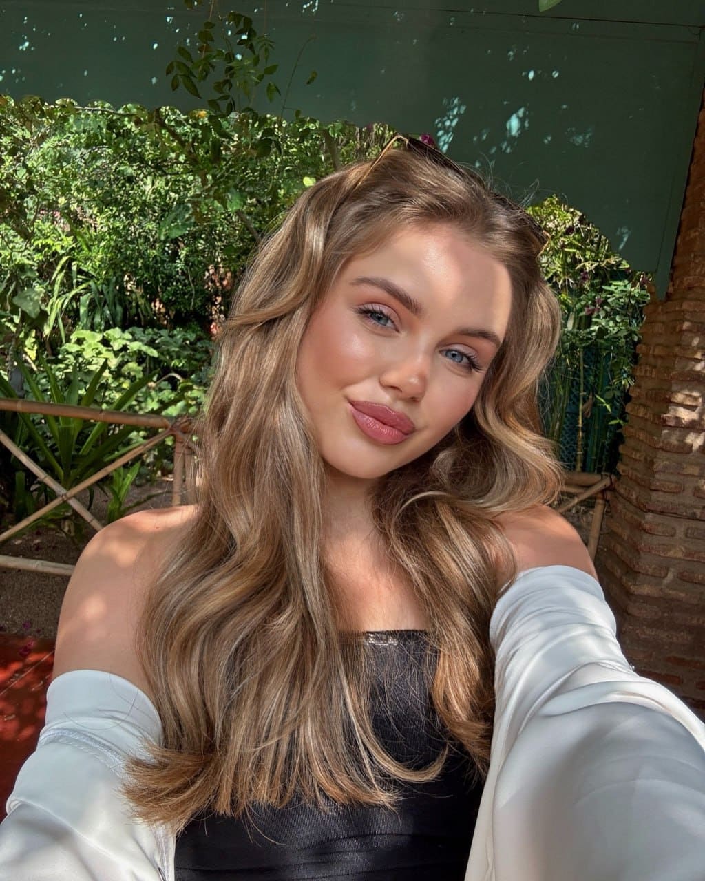 Selfie of a woman in a black top and white off-the-shoulder jacket, with foliage and a brick wall behind.