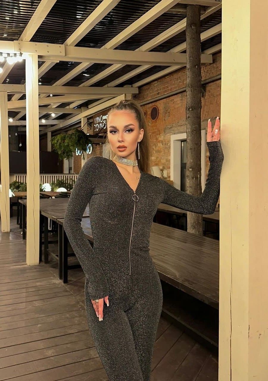A woman with hair tied back stands on a wooden deck in a sparkly dark gray jumpsuit, hand resting on a post.