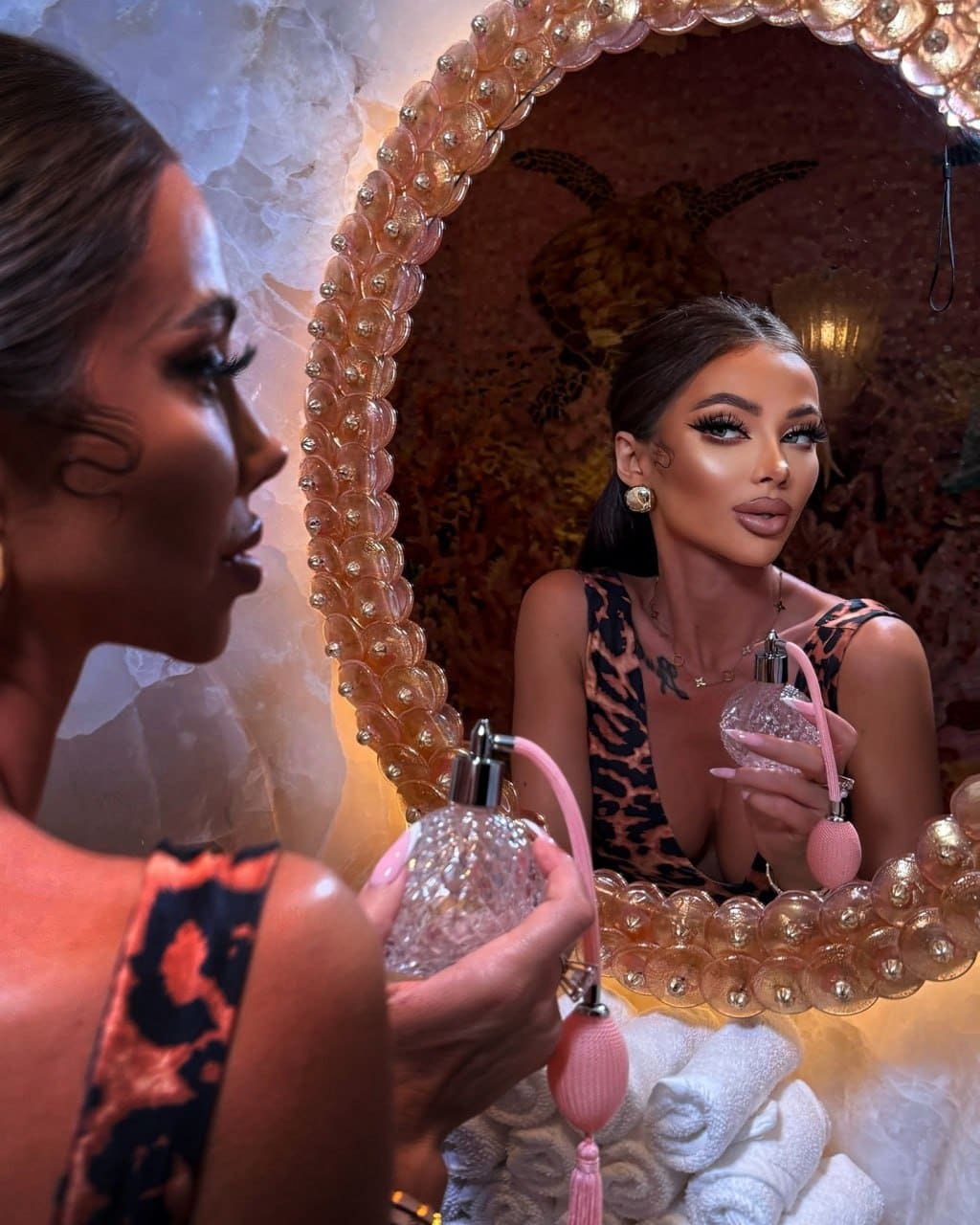 A woman in profile holds a pink perfume bottle, while her mirror reflection shows her facing forward in a leopard-print top with a deep neckline.